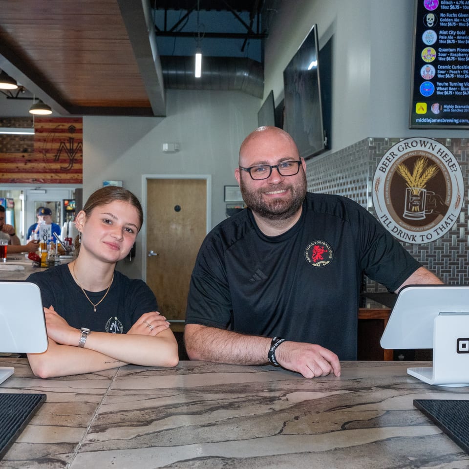 Two smiling staff members pose behind a brewery bar with registers, beer taps, and a digital beer menu.