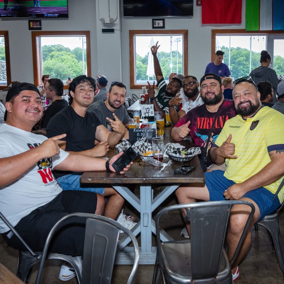 Friends pose together around a restaurant table with drinks and food, while sports play on televisions and flags decorate the room.